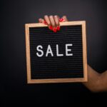 Close-up of a hand with red nails holding a SALE sign on black felt board.