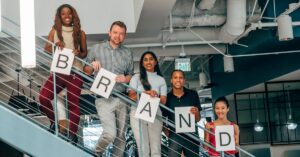 Diverse group of adults smiling and holding 'BRAND' signs on an office staircase.